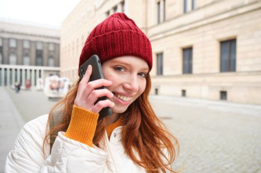 Mobile broadband and people. Smiling young redhead woman walks in town and talks on mobile phone, calling friend on smartphone, using internet to make a call abroad.