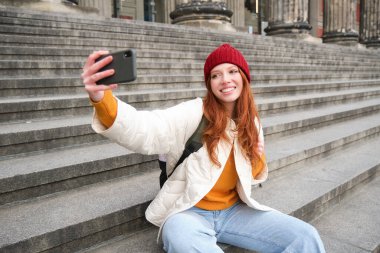 Stylish young girl in red hat, takes photos on smartphone camera, makes selfie as she sits on stairs near museum, posing for photo with app filter.