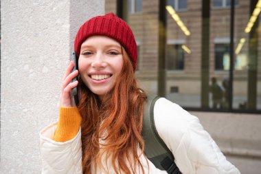 Young people and mobile connection. Happy redhead girl talks on phone, makes telephone call, stands outdoors with backpack and uses smartphone app.