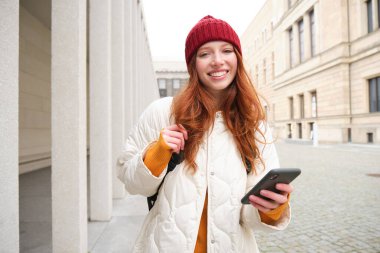 Tourism and technology. Beautiful redhead girl tourist, looking for a route on application, using city map on smartphone, reading about sightseeing places on mobile phone.