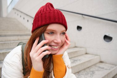 Young stylish redhead girl in red hat, sits on street and talks on mobile phone, has telephone conversation, rings her friend while relaxes outdoors.