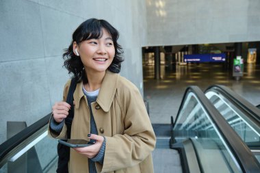 Telsiz kulaklıklı esmer kızın portresi, müzik dinliyor, akıllı telefon kullanıyor, yürüyen merdivene çıkıyor. Boşluğu kopyala