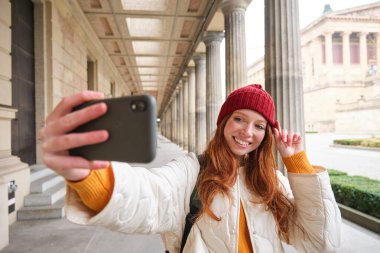 Havalı kızıl saçlı turist, turizmin önünde selfie çekiyor, akıllı telefonla fotoğraf çekiyor, mobil kameraya bakıyor ve poz veriyor..