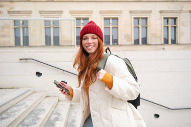 Smiling redhead girl, student tourist walks around city, goes up the stairs, looks at mobile phone map to get around town, sends message on smartphone.