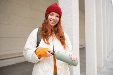 Beautiful redhead woman drinking hot tea or coffee from thermos, female tourist enjoys warm drink, rests during her journey around town.