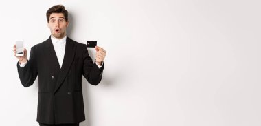 Siyah takım elbiseli yakışıklı iş adamının görüntüsü. Şaşırmış görünüyor ve beyaz arka planda duran cep telefonu ekranıyla kredi kartını gösteriyor..