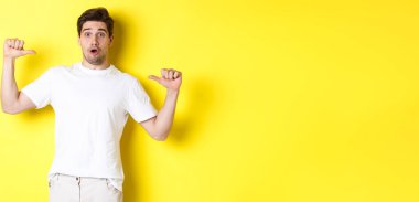 Şaşkın yakışıklı adam kendini işaret ediyor, şaşkın şaşkın bakıyor, sarı arka planda duruyor. Boşluğu kopyala