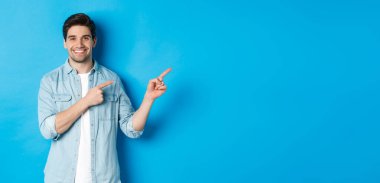 Çekici yetişkin bir adamın portresi gülümsüyor, parmaklarını logoya ya ya da afişe doğrultuyor, mavi arka plana karşı reklam gösteriyor..