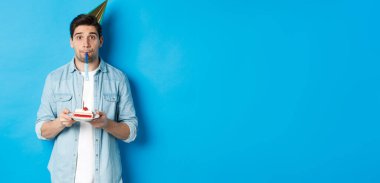 Doğum günü pastası tutan yakışıklı genç adam, parti şapkası takıyor ve düdük çalıyor, mavi arka planda duruyor..