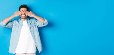 Gülümseyen yetişkin bir adam şaşkınlık bekliyor, elleriyle gözlerini kapatıyor ve mavi arka planda günlük kıyafetlerle dikiliyor..