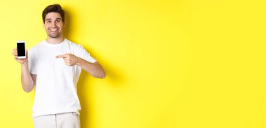 Akıllı telefon ekranını işaret eden çekici bir genç adam görüntüsü, bir uygulama gösteriyor, sarı arka planda duruyor..