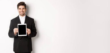 Güzel takım elbiseli yakışıklı işadamının portresi, dijital tablet ekranı gösteriyor ve gülümsüyor, beyaz arka planda duruyor..