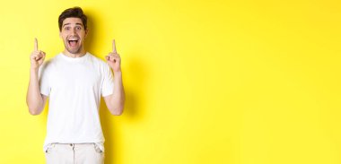 Beyaz tişörtlü heyecanlı yakışıklı adamın portresi, parmakları havada, teklifini gösteriyor, sarı arka planda duruyor..