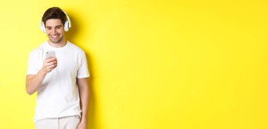 Kulaklıklı genç adam akıllı telefondan mesaj okuyor, gülümsüyor, kulaklıkla müzik dinliyor ve uygulamadan şarkı seçiyor, sarı arka planda duruyor..