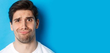 Şüpheci ve hayal kırıklığına uğramış bir adamın resmi. Karmaşık suratlı, mavi arka planda duruyor..