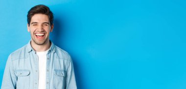 Mutlu, genç, sakallı, sıradan kıyafetler giymiş, gülen, neşeli, mavi arka planda duran bir adamın yakın çekimi..