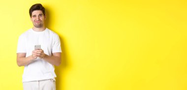 Hoşnutsuz ve isteksiz bir adam suratını buruşturuyor, akıllı telefondaki mesajdan rahatsız oluyor, sarı arka planda duruyor..