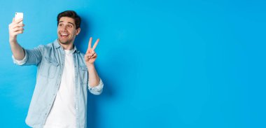 Mutlu adam selfie çekiyor ve mavi arka planda barış işareti gösteriyor, elinde cep telefonu var..