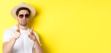 Turizm ve tatil kavramı. Yazlık şapkalı, güneş gözlüklü, kameraya silah doğrultan, sarı arka planda duran havalı adamın yakın çekimi..