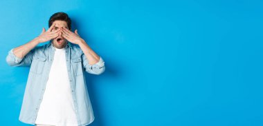 Şok olmuş bir adam gözlerini kapatıyor ve parmaklarının arasından bakıyor. Utanç verici bir şeye bakıyor. Mavi arka planda duruyor..