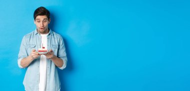 Doğum günü. Şaşkın adam doğum günü pastasına bakıyor, mavi arka planda duruyor..