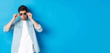 Kendinden emin maço adam güneş gözlüğü taktı, havalı ve küstah görünüyordu, mavi arka planda duruyordu..