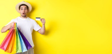 Ürün ve kredi kartıyla alışveriş torbaları gösteren etkileyici bir adam, sarı arka planda duruyor..