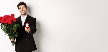 Siyah takım elbiseli yakışıklı, elinde güller ve nişan yüzüğü olan, onunla evlenme teklifi eden, beyaz arka planda duran....