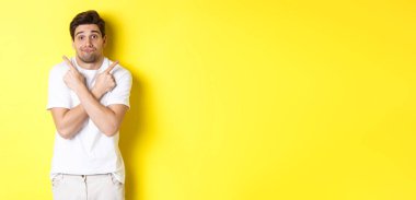 Kararsız adam parmaklarını yana çeviriyor, karar vermek için çabalıyor, tavsiye istiyor, sarı arka planda duruyor..