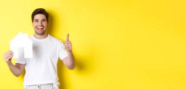 Emlak konsepti. Mutlu genç adam kağıt ev modelini gösteriyor ve onaylıyor, komisyoncuyu tavsiye ediyor, sarı arka planda duruyor..