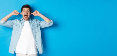 Sinirli adam, gürültücü komşuların kulaklarını kapatır, yukarı bakar ve bağırır, yüksek sesli müzik duyar, mavi arka planda durur..