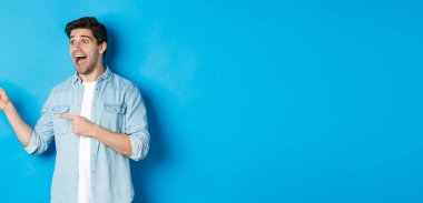 Yakışıklı bir adamın portresi. Mavi arka planda dikilip, heyecanla sol parmaklarını işaret ediyor..