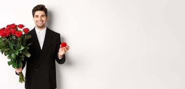 Siyah takım elbiseli yakışıklı bir adam, elinde bir buket kırmızı gül ve yüzük, evlenme teklif ediyor, kendinden emin gülümsüyor, beyaz arka planda duruyor..