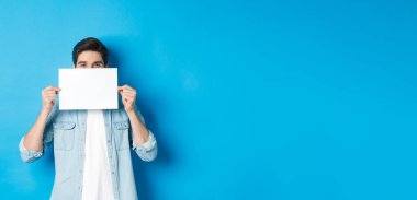 Sinsi yakışıklı adam logon için boş bir kağıt parçasının arkasına saklanıyor, duyuru yapıyor ya da tanıtım teklifi yapıyor, mavi arka planda duruyor..