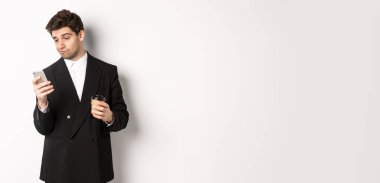 Düşünceli yakışıklı iş adamının portresi, kahve içen ve internette gezinen, akıllı telefon ekranına bakan, beyaz arka planda duran.