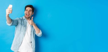 Akıllı telefondan selfie çeken yakışıklı ve hareket eden kameraya silah doğrultan arsız, mavi arka planda duran....