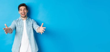 Dost canlısı bir adam elini uzatıyor ve seni selamlıyor, misafirleri selamlıyor ya da sarılıyor, mavi arka planda duruyor..