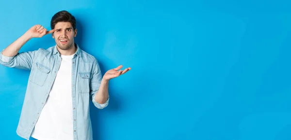 Confused man scolding person for being stupid, pointing finger at head and shrugging, looking puzzled, disapprove actions, standing against blue background.
