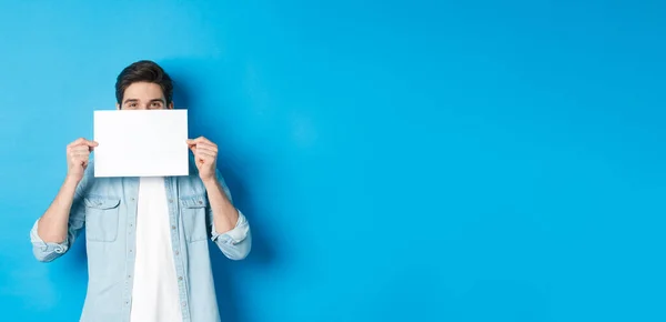 Sneeky handsome guy hiding face behind blank piece of paper for your logo, making announcement or showing promo offer, standing over blue background.