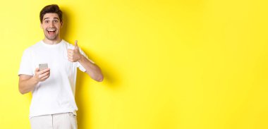 Akıllı telefonu tutan mutlu bir adam, onaylanmış bir şekilde baş parmağını gösteriyor, sarı arka planda duran, online bir şey öneriyor..