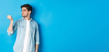 Gözlüklü genç adam tırnaklarına bakıyor, maniküre bakıyor, mavi arka planda duruyor..