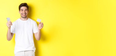 Alışveriş yapmayı düşünen mutlu bir adam, elinde kredi kartı ve akıllı telefon tutan, gülümseyen, sarı arka planda duran,.