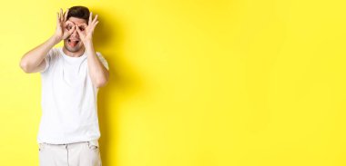 Genç adam komik yüzler ve yapışkan dil gösteriyor, sarı arka planda şakacı duruyor..