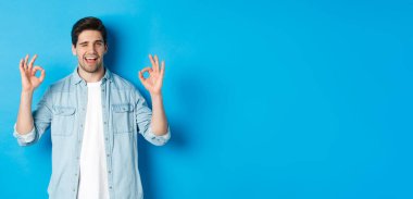 Rahat ve kendinden emin bir adam tamam işaretler ve göz kırpma, her şey yolunda jest, mavi arka planda duruyor..