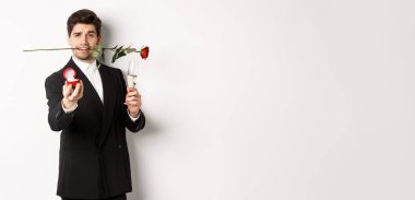 Takım elbiseli romantik genç bir adam evlenme teklif ediyor, elinde gül, bir kadeh şampanya, nişan yüzüğünü gösteriyor, evlenme teklif ediyor, beyaz arka planda duruyor..