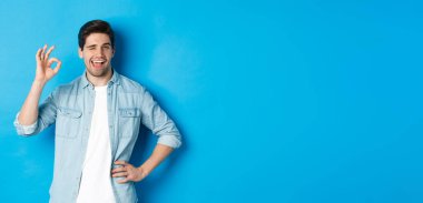 Kendine güveni tam bir yetişkin adam göz kırpıyor ve tamam işareti gösteriyor, iyi bir şey onayla, mavi arka planda duruyor..