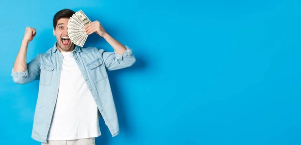 Successful young man earn money, making fist pump and showing cash, winning prize or receive credit, standing over blue background.