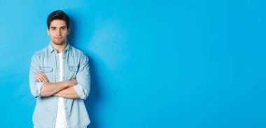 Sıradan giyinmiş, ciddi ve kendinden emin, mavi arka planda duran yakışıklı beyaz bir adam resmi..