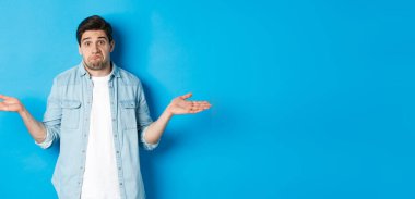 Kafası karışmış genç adam omuzlarını silkiyor, hiçbir şey bilmiyor, habersiz görünüyor, mavi arka planda duruyor..