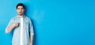 Gergin adam kendini işaret ediyor ve kafası karışık görünüyor. Mavi arka plan üzerinde sıradan kıyafetler giyiyor..